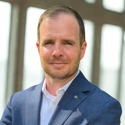TJ Alexander, digital marketing consultant and founder of TJA Strategies, wearing a blue suit and standing in front of large windows with a confident expression.