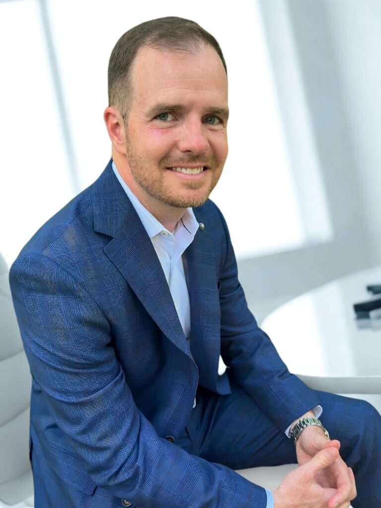 TJ Alexander, founder of TJA Strategies, seated in a modern office setting, wearing a blue suit and smiling confidently.